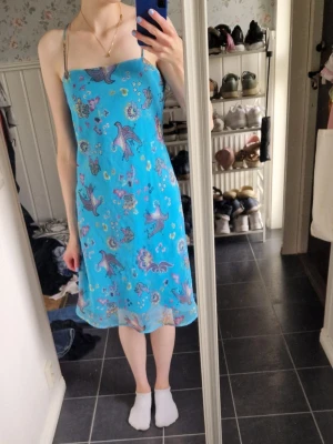 Blå klänning  - Supersöt blå vintage klänning med tunna axelband och blommigt mönster i lila, rosa och gult. Klänningen har en rak passform och är knälång, perfekt för varma dagar. Materialet känns lätt och luftigt, vilket gör den skön att bära under sommaren.