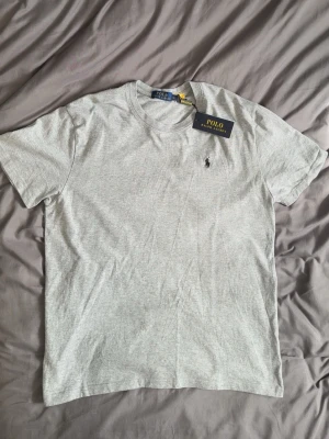 Grå t-shirt från Polo Ralph Lauren - Klassisk grå t-shirt från Polo Ralph Lauren med rund halsringning och korta ärmar. T-shirten har den ikoniska broderade loggan på bröstet och är tillverkad i mjuk bomull för en skön känsla. Perfekt basplagg till garderoben.