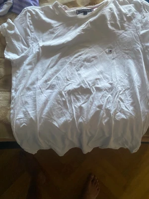 Vit t-shirt från Tommy Hilfiger - Klassisk vit t-shirt från Tommy Hilfiger med liten broderad logga på bröstet. T-shirten har rund hals och korta ärmar. Tillverkad i mjuk bomull som känns skön mot huden. Perfekt basplagg för alla tillfällen.