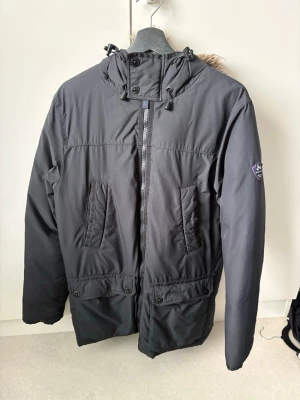 Svart parkas från Jean Paul - Snygg svart parkas från Jean Paul med avtagbar huva och dragsko. Jackan har två stora fickor med knappar, två snedställda bröstfickor och ett emblem på ärmen. Perfekt för kalla dagar med sin klassiska design och praktiska detaljer.