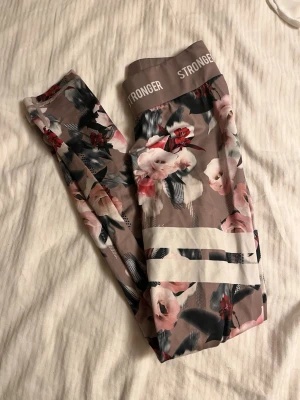 Blommiga leggings från Stronger -  Leggings från Stronger med blommigt mönster i rosa, rött, vitt och svart på beige botten. Säljer då de inte kommer till användning, använt fåtal gånger.
