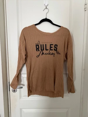 Beige långärmad tröja med tryck - Trendig beige långärmad tröja med texten 'RULES maker' och fjärilar framtill. Tillverkad i ett ribbat, mjukt material med avslappnad passform och rund halsringning. Perfekt för en avslappnad och cool stil.