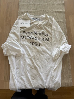 Vit Acne Studios t-shirt - Vit t-shirt från Acne Studios med tryck 'Acne Studios STOCKHOLM 1996' på bröstet. Klassisk passform och rund halsringning. Perfekt basplagg för en clean och stilren look.