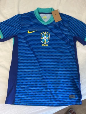 Blå Brasilien fotbollströja Nike - Säljer en blå Brasilien fotbollströja från Nike med CBF-emblem och fem stjärnor på bröstet. Tröjan har tunna horisontella ränder i olika blå nyanser, gula detaljer och ljusblå krage. Perfekt för dig som älskar fotboll och vill stötta Brasilien.