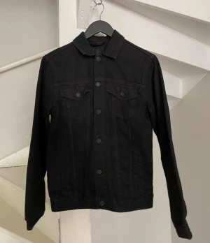 Svart klassisk jeansjacka - Stilren svart jeansjacka med raka linjer och klassisk krage. Jackan har två bröstfickor med lock och knappar framtill. Perfekt basplagg för dig som gillar en enkel men snygg look. Långa ärmar och knappstängning hela vägen.