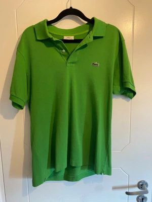 Grön pikétröja från Lacoste - Snygg grön pikétröja från Lacoste med klassisk krage och korta ärmar. Tröjan har den ikoniska krokodilloggan broderad på bröstet och är tillverkad i bomull för en bekväm känsla. Perfekt för dig som gillar stilrena och sportiga plagg. Storlek 3/M