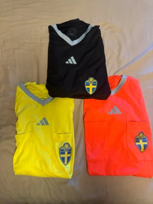 Domartröjor Adidas Sverige - Tre domartröjor från Adidas i svart, gul och neonorange med grå V-ringning. Alla har Svenska Fotbollförbundets märke på bröstet och två bröstfickor. Perfekt för dig som dömer fotbollsmatcher och vill ha officiell look.