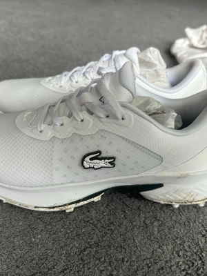 Vita sneakers från Lacoste golfskor - Snygga vita sneakers från Lacoste med klassisk krokodillogo på sidan. Skorna har lågt skaft, vita snören och ovandel i mesh och syntetmaterial för en sportig look. Perfekta för dig som gillar stilrena och fräscha sneakers.