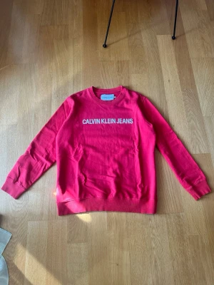 Röd sweatshirt från Calvin Klein Jeans - Röd sweatshirt från Calvin Klein Jeans med vit logga tryckt på bröstet. Klassisk rund halsringning, ribbade muddar vid ärmslut och nederkant. Perfekt för en avslappnad och trendig look. Storlek xl men kan även sitta som en L.