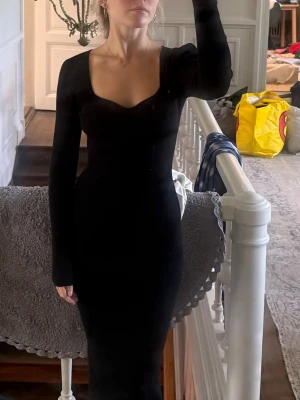 Adoore klänning - Snygg svart långklänning med lång ärm och ribbad struktur. Klänningen har en djup v-ringning och sitter tight mot kroppen för en stilren och modern look. Perfekt för dig som gillar minimalistisk stil.