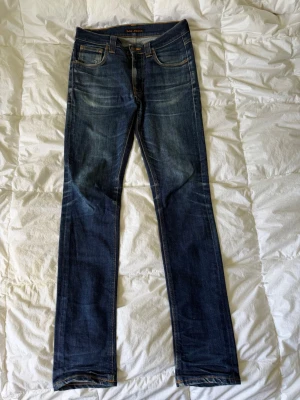 Mörkblå skinny jeans från Nudie Jeans W30/L34 - Säljer ett par mörkblå skinny jeans från Nudie Jeans med klassiska fem fickor och snygga slitningar. Jeansen har kontrastsömmar i orange och är tillverkade i stretchig denim för en bekväm passform. Perfekta för dig som gillar en smal siluett.