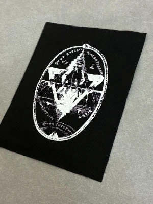 Svart tygpatch med mystiskt tryck - Svart tygpatch med vitt tryck i form av en oval med geometriska symboler och latinsk text. Motivet har en alternativ och mystisk känsla, perfekt att sy fast på jacka eller väska för en personlig stil.