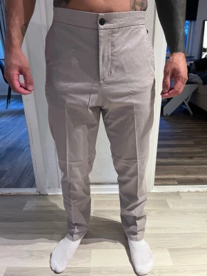 Beige kostymbyxor med rak passform - Snygga beige kostymbyxor med rak passform och pressveck. Byxorna har elastisk midja baktill, två bakfickor och stängs med knapp och dragkedja. Perfekta för en stilren look och lätta att matcha med skjorta eller t-shirt.