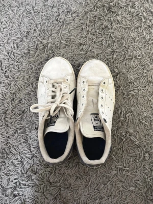 Vita Adidas Stan Smith sneakers - Klassiska vita Adidas Stan Smith sneakers med mörkblå detalj på hälen och perforerade ränder på sidorna. 