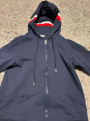 Mörkblå Moncler hoodie med dragkedja - Snygg mörkblå hoodie från Moncler med dragkedja framtill och klassisk Moncler-logga på ärmen. Huva med snörning och ribbade muddar i rött, vitt och svart. Perfekt för en avslappnad och trendig look. Tillverkad i mjukt bomullsmaterial.