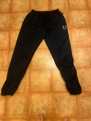 Svarta träningsbyxor Under Armour XXL - Säljer ett par svarta mjukisbyxor från Under Armour med resår i midjan och vid bensluten. Byxorna har en avslappnad passform och diskret logga på vänster lår. Perfekta för chill eller träning.