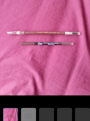 Bruna ögonbrynspennor med borste - Två ögonbrynspennor i bruna nyanser, båda med inbyggd borste i ena änden. Ena pennan har guldtext och transparent hylsa, den andra har svart text och heter 'Smoky Slim Brow Pencil'. Perfekt för att forma och fylla i brynen med precision.