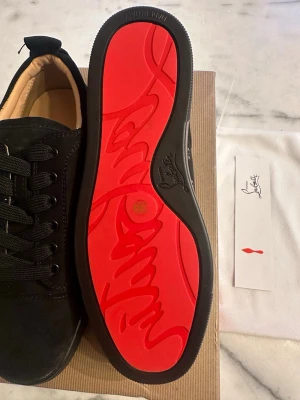 Svarta sneakers med röd sula - Stilrena svarta sneakers från Christian Louboutin med ikonisk röd sula och svarta skosnören. Ovandelen är i mocka och insidan är beige. Perfekta för dig som vill ha en lyxig och trendig look.