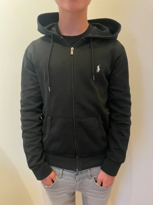 Svart zip hoodie från Polo Ralph Lauren - Säljer en svart hoodie med dragkedja från Polo Ralph Lauren. Klassisk design med vit logga på bröstet, huva med dragsko och kängurufickor framtill. Perfekt till jeans och enkel att matcha. Mjuk bomullskänsla och ribbade muddar. Storlek S