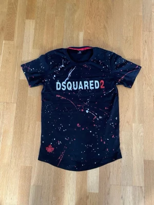 Svart Dsquared2 t-shirt med färgstänk - Snygg svart t-shirt från Dsquared2 med coola färgstänk i vitt och rött samt stor logga framtill. T-shirten har rund hals, korta ärmar och en relaxed passform. Perfekt för dig som vill sticka ut med en unik och trendig look.