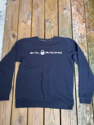 Mörkblå Sail Racing sweatshirt - Mörkblå sweatshirt från Sail Racing med tryckt logga på bröstet. Klassisk rund halsringning och ribbade muddar vid ärmslut och nederkant. Perfekt för en avslappnad och sportig stil.