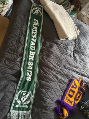 Grön Färjestad BK halsduk - Säljer en lång, grön halsduk med vita detaljer och texten 'Färjestad BK 24/25' samt klubbens logga och Karlstad. Halsduken har vita fransar i ändarna och är perfekt för att visa ditt stöd på matchen.