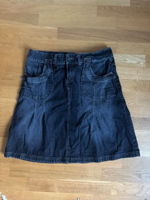Svart jeanskjol med fickor - Cool svart jeanskjol med stora fram- och bakfickor samt bälteshällor. Kjolen har en klassisk jeanslook med synliga sömmar och knappstängning framtill. Perfekt för en avslappnad och trendig stil.