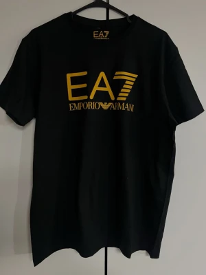 Svart EA7 Emporio Armani t-shirt - Snygg svart t-shirt från EA7 Emporio Armani med stort gult tryck framtill. Klassisk rund halsringning och korta ärmar. Perfekt för dig som gillar stilrena och trendiga plagg med tydlig logga. Tillverkad i mjuk bomull för skön känsla.