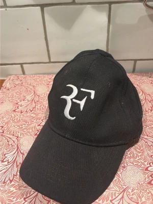 Svart keps med vit RF-brodyr - Säljer brosans keps, Snygg svart keps med böjd skärm och vit broderad RF-logga framtill. Klassisk modell med justerbar rem bak. Perfekt för dig som vill ha en stilren accessoar med sportig känsla.