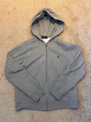  polo rl zip hoodie - Snygg grå hoodie med dragkedja från Polo Ralph Lauren. Klassisk design med huva, två fickor framtill och den ikoniska lilla loggan broderad på bröstet. Tillverkad i mjukt sweatshirtmaterial som är skönt att ha på sig. Perfekt till chill dagar eller när du vill vara casual.