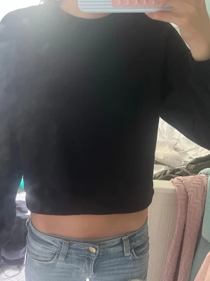Svart croppad hoodie - Säljer en svart croppad hoodie med rund hals och långa ärmar. Hoodien har en enkel och clean design utan tryck eller detaljer, perfekt till jeans eller mjukisbyxor. Materialet ser ut att vara mjukt och bekvämt, troligen bomullsblandning.