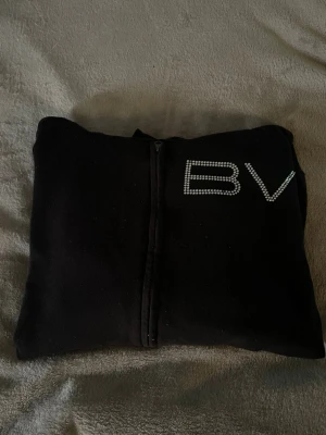 Svart hoodie med dragkedja och BV-detalj - Säljer en svart hoodie med dragkedja och huva. På bröstet finns BV-bokstäver i glittrande stenar som ger en snygg touch. Hoodien har fickor framtill och ribbade muddar vid ärmar och nederkant. Perfekt för en chill och stilren look.