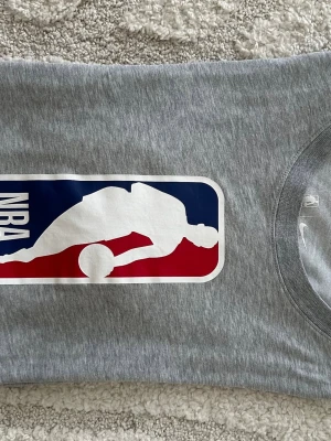 Grå NBA Nike Dri-FIT t-shirt - Säljer en grå t-shirt från Nike med stor NBA-logga framtill. Tillverkad i mjukt Dri-FIT-material som andas, perfekt för dig som gillar basket eller streetwear. Klassisk passform och rund halsringning.