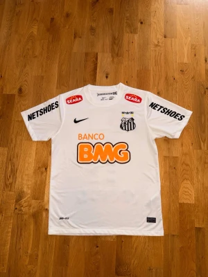 Vit Santos fotbollströja Neymar Jr - Säljer en vit Santos fotbollströja från Nike med Neymar Jr och nummer 11 på ryggen. Tröjan har tryckta sponsorer som BMG, Netshoes och Seara samt klubbmärke på bröstet. Tillverkad i lätt och ventilerande material, perfekt för träning eller match.