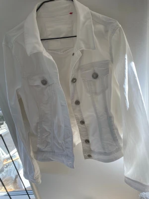 Vit klassisk jeansjacka - Snygg vit jeansjacka med silverfärgade knappar och två bröstfickor med lock, två innerfickor och fejkfickor på sida. Jackan har en tidlös design med krage och raka ärmar. Perfekt att slänga över en t-shirt för en fräsch look.