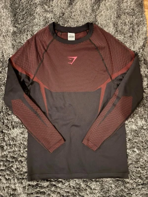 Svart och vinröd långärmad träningströja - Snygg långärmad träningströja från Gymshark i svart och vinrött. Tröjan har ventilerande meshpaneler och tight passform, perfekt för gym eller löpning. Materialet är stretchigt och följsamt för maximal rörelsefrihet. Diskret logga på bröstet. Kontakta vid intresse!