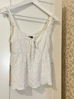Vit linnetopp - spets - Söt vit linnetopp med smala spetsband och knyt framtill. Toppen har en rynkad struktur och är ärmlös, perfekt för varma dagar. Passar till jeans eller kjol för en avslappnad och trendig look.