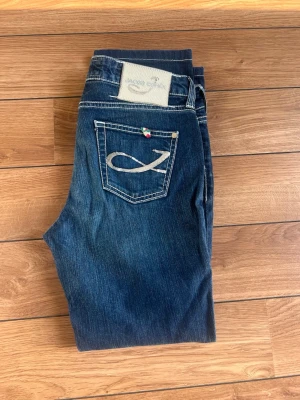 Mörkblå jeans med broderad ficka - Snygga mörkblå jeans med kontrastsömmar och broderad detalj på bakfickan. Klassisk femficksmodell med knapp- och dragkedjegylf. Jeansen har en lätt tvättad look och raka ben. Perfekta för en avslappnad och trendig stil.