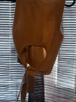 Brun stickad väst med knytband - Säljer en brun stickad väst i ribbat material. Västen är ärmlös med rund halsringning och har snygga knytband i midjan som ger en cool detalj. Perfekt att styla över en skjorta eller t-shirt för en trendig lager-på-lager-look.