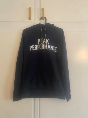 Mörkblå Peak Performance hoodie - Snygg mörkblå hoodie från Peak Performance med vit logga på bröstet. Klassisk modell med huva och dragsnören samt en stor magficka framtill. Perfekt för dig som gillar en enkel och stilren look. Materialet känns mjukt och bekvämt.