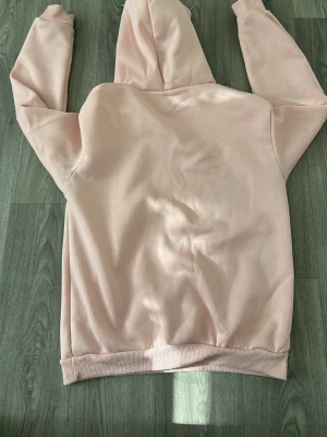 Ljusrosa hoodie med magficka - En stilren ljusrosa hoodie med huva och dragsnören. Hoodien har en klassisk magficka framtill och ribbade muddar vid ärmslut och nederkant. Perfekt för en avslappnad och trendig look. Materialet känns mjukt och bekvämt mot huden.