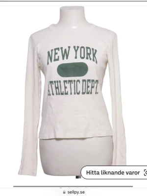 Vit långärmad topp med tryck - Vit långärmad topp med grönt tryck på bröstet där det står 'NEW YORK ATHLETIC DEPT.'. Rund halsringning och rak passform. Perfekt för en avslappnad och sportig stil.