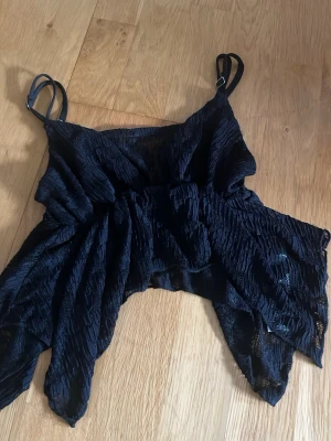 Svart omlott topp från SHEIN - Säljer en svart omlott topp från SHEIN med tunna axelband och asymmetrisk nederkant. Tyget är lätt och har en snygg struktur som ger toppen en unik look. Perfekt för dig som vill ha något trendigt och annorlunda i garderoben.