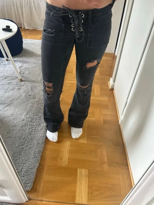 Svarta bootcut jeans från Gina tricot - Säljer ett par svarta bootcut jeans med snörning i midjan och slitna detaljer på låren och knäna. Jeansen har fickor fram och bak samt en cool, trendig look som passar perfekt till streetwear-stilen.