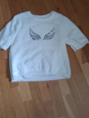 Vit fluffig stickad tröja Kappahl - Supersöt vit stickad tröja från Kappahl i fluffigt material. Tröjan har ett par silvriga paljettvingar på bröstet som ger en unik och trendig look. Perfekt för dig som gillar att sticka ut lite extra med din outfit.