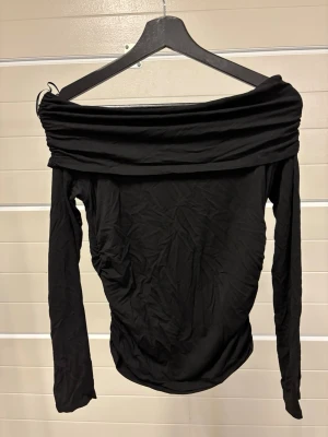 Svart offshoulder topp Gina Tricot - Trendig svart offshoulder topp från Gina Tricot med långa ärmar och bred krage som vilar över axlarna. Tillverkad i mjukt material som sitter snyggt och markerar siluetten. Perfekt för dig som vill ha en stilren och modern look.