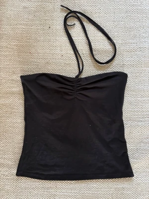 Svart linne med knytband - Säljer ett svart linne med smala knytband i nacken och rynkdetalj framtill. Toppen är ärmlös och har en enkel, stilren design som passar till det mesta. Perfekt för dig som gillar minimalistisk stil.