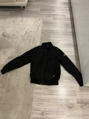Svart vindjacka med hög krage - Storleken är xl men passar L bättre, Snygg svart vindjacka med hög krage och dragkedja framtill. Jackan har ribbade muddar vid ärmslut och nederkant samt en patch på vänster ärm. Materialet ser ut att vara lätt och vindtätt, perfekt för blåsiga dagar.