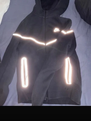 Svart Nike hoodie med reflexdetaljer - Svart hoodie från Nike med dragkedja och huva. Snygga vita reflexdetaljer längs bröstet och vid fickorna. Perfekt för dig som vill sticka ut lite extra. Klassisk Nike-logga på bröstet och två fickor framtill. Skön och sportig stil.