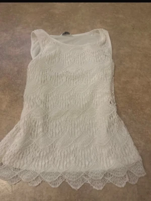 Vit ärmlös spetslinne - Säljer ett vitt ärmlöst linne med vacker spets och vågigt mönster. Linnet har rund halsringning och spetsdetaljer längs nederkanten. Perfekt att styla med jeans eller kjol för en fräsch look.